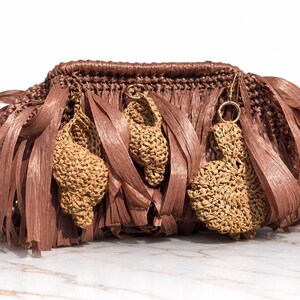 May include: A brown crocheted clutch bag featuring long, flat fringe strips and three dangling, textured, light tan crocheted shell-shaped charms attached to the front with small gold-toned metal rings. The bag has a structured, woven top edge.