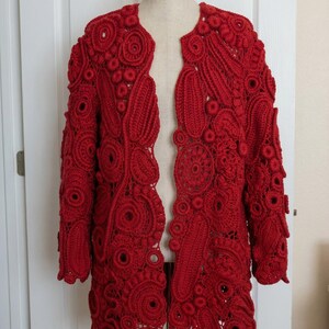 May include: A vibrant red, open-front crochet jacket displayed on a mannequin. The jacket features an intricate, all-over pattern of circular and floral motifs. The sleeves are long, and the overall design is eye-catching and detailed, showcasing the craftsmanship.