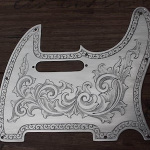 Peut inclure: Un médiator de guitare en métal argenté orné d'un motif gravé complexe en volutes sur toute sa surface. Le design comprend des motifs détaillés en forme de feuilles et une bordure décorative, avec des trous de vis et une découpe pour micro.