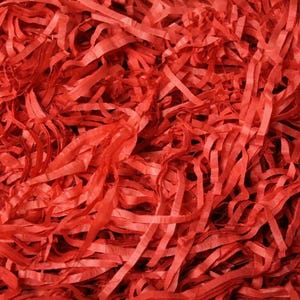 May include: A dense collection of vibrant red shredded paper strips. The material consists of thin, crinkled, and intertwined ribbons of paper that create a textured, layered surface with a consistent bright red color throughout the entire display.