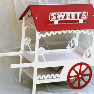 May include: A white and red wooden sweets cart featuring a roof with the word SWEETS in white lettering. The cart has a scalloped trim, two wheels, and decorative cutouts of lollipops and candies on the side panels and frame structure.
