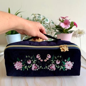 May include: A navy blue velvet cosmetic bag with a gold zipper and handle. The bag features an embroidered floral design in pink and green. The zipper pull is a gold flower. The bag is held by a hand.