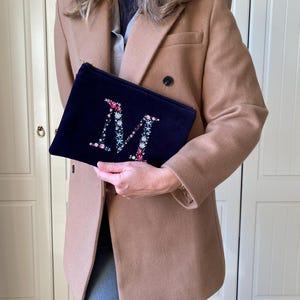 May include: A navy blue velvet clutch with a floral embroidered letter "M" in pink, white, and green.