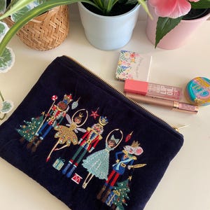 Puede incluir: Una bolsa de terciopelo azul oscuro con cremallera y un diseño bordado de un Cascanueces y una bailarina. El diseño incluye un árbol de Navidad, regalos y otros detalles festivos.