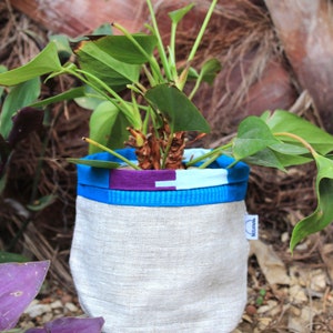 Peut inclure: Un pot de plante en tissu avec un extérieur gris clair et un bord à motifs bleu, violet et bleu clair. Le pot contient une plante verte avec de grandes feuilles. Le pot est posé sur une pierre.