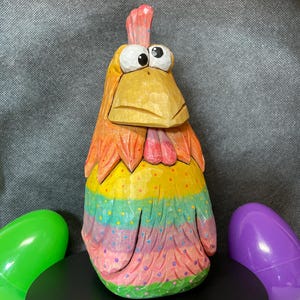 May include: A hand-carved wooden chicken figurine with a colorful rainbow paint scheme. The chicken has large, googly eyes and a pink comb.