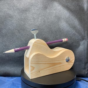 May include: A wooden pencil holder with a pencil inserted. The holder is made of light-colored wood and has a curved design. A purple pencil is inserted through the holder. A metal screw is on top of the holder.