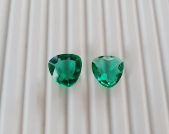 Op de afbeelding: Twee gefacetteerde, smaragdgroene edelstenen met een driehoekige vorm. De edelstenen worden gepresenteerd op een wit, geribbeld oppervlak, dat hun helderheid en glans benadrukt. De edelstenen zijn waarschijnlijk bedoeld voor het maken van sieraden.