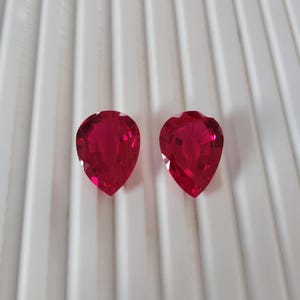 May include: Two faceted, pear-shaped, bright red gemstones. The gems are displayed against a white, ridged surface. The gems have a high degree of clarity and brilliance, suggesting they are of high quality. Ideal for jewelry making.