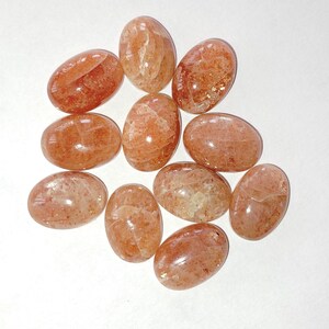 Puede incluir: Una colección de piedras preciosas de sol en forma ovalada. Las piedras tienen un tono cálido, naranja-rojizo, con inclusiones brillantes. Las piedras pulidas son lisas y varían ligeramente en tamaño y color, mostrando variaciones naturales.