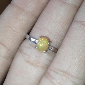 Könnte beinhalten: Ein silberner Ring mit einem ovalen Opal-Edelstein. Der Opal ist hellgelb mit innerem Schimmern. Das dünne, polierte Band wird von vier Krappen gehalten. Ein klassisches Schmuckstück.