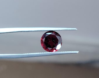 Pode incluir: Uma gema redonda, facetada, de cor vermelha escura é segurada por pinças prateadas contra um fundo desfocado. O corte da gema permite que a luz se reflita, criando um efeito brilhante. A gema parece ser uma rubi ou uma granada.