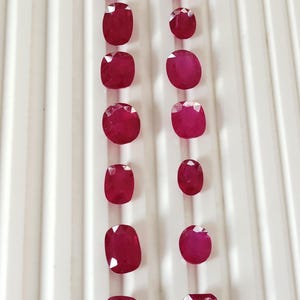 Pode incluir: Uma coleção de rubis facetados, vermelho escuro, em várias formas, incluindo cortes redondos, ovais e almofada. As pedras preciosas são dispostas em fileiras verticais sobre um fundo branco, mostrando suas superfícies polidas e sua cor vibrante.