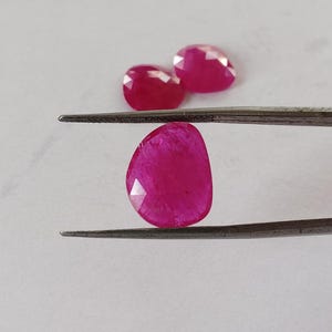 Puede incluir: Una gema de rubí rosa oscuro facetada es sostenida por unas pinzas, con dos rubíes adicionales en el fondo. Las gemas tienen una superficie pulida y reflectante, sobre un fondo blanco.