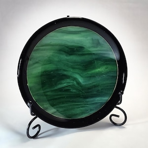May include: A round green glass plate with a black rim, displayed on a black metal stand. The glass has a swirling, marbled pattern.