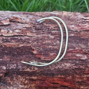 Puede incluir: Una pulsera abierta de color dorado con un diseño de doble banda. La pulsera tiene una forma curva con un detalle en espiral en un extremo. Los extremos de la pulsera tienen pequeñas puntas de color claro. La pulsera se muestra sobre una superficie marrón texturizada.