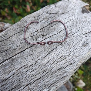 May include: A dark copper-colored metal cuff bracelet with a curved, open design. The bracelet features two rounded, looped ends that face each other in the center, creating a symmetrical, minimalist aesthetic with a smooth, polished finish.