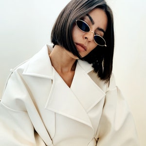 May include: A woman wearing a white, belted trench coat with a long, straight, dark brown bob hairstyle. She is wearing oval-shaped sunglasses with a gold frame.