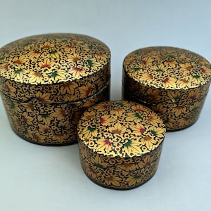 May include: Three round decorative boxes with intricate floral patterns in gold, green, and red on a black background. The boxes are made of papier-mâché and are nested inside each other.
