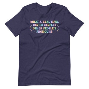 May include: A navy blue t-shirt with the text "WHAT A BEAUTIFUL DAY TO RESPECT OTHER PEOPLE'S PRONOUNS" in rainbow colors with glitter accents.