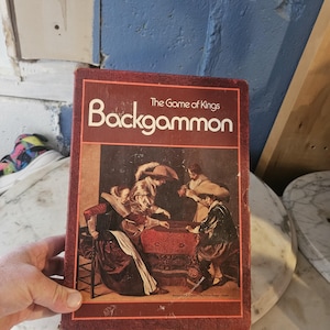 Puede incluir: Una caja de juego de Backgammon vintage con el título "The Game of Kings". La portada presenta una pintura de personas jugando al juego. La caja es de color rojo oscuro con texto blanco.