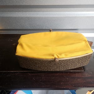 May include: A vintage, rectangular basket with a woven brown base and a bright yellow fabric top. The basket has a wooden frame and a small, round clasp. The item is sitting on a dark wooden surface.