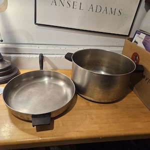 May include: A stainless steel frying pan and a pot with black handles sit on a wooden surface. The pan is round with a single handle, while the pot is cylindrical. The background includes a framed print and a lamp.