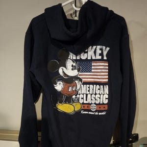 Puede incluir: Sudadera con capucha azul marino con un gr&aacute;fico de Mickey Mouse, la bandera estadounidense y las palabras "MICKEY AMERICAN CLASSIC". Tiene una capucha con cord&oacute;n y pu&ntilde;os y dobladillo acanalados.