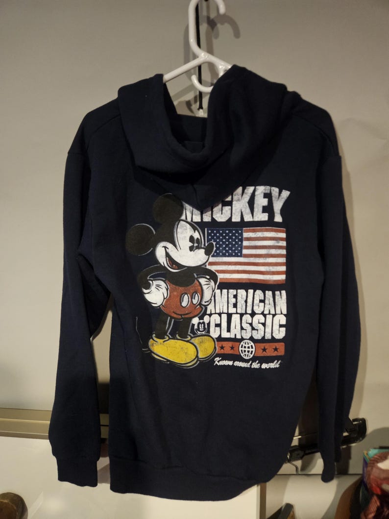 Puede incluir: Sudadera con capucha azul marino con un gr&aacute;fico de Mickey Mouse, la bandera estadounidense y las palabras "MICKEY AMERICAN CLASSIC". Tiene una capucha con cord&oacute;n y pu&ntilde;os y dobladillo acanalados.