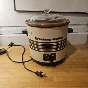 May include: Cream-colored Crockery-Kettle slow cooker with brown stripes and a glass lid. The appliance has a black control knob and handles. The power cord is plugged in. The text "Crockery-Kettle" is printed on the front.