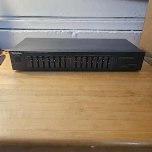 Puede incluir: Un ecualizador gráfico Technics SH-8050 negro con múltiples deslizadores y luces rojas iluminadas. El dispositivo está sobre una superficie de madera. El logotipo de Technics es visible en el lado izquierdo.