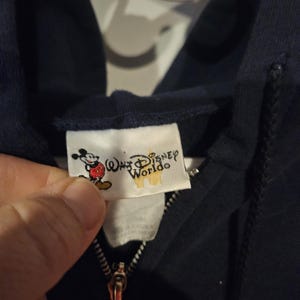 Puede incluir: Sudadera con capucha azul marino con cremallera y una etiqueta con Mickey Mouse y las palabras "Walt Disney World". La cremallera es dorada. La sudadera est&aacute; hecha de un material suave.