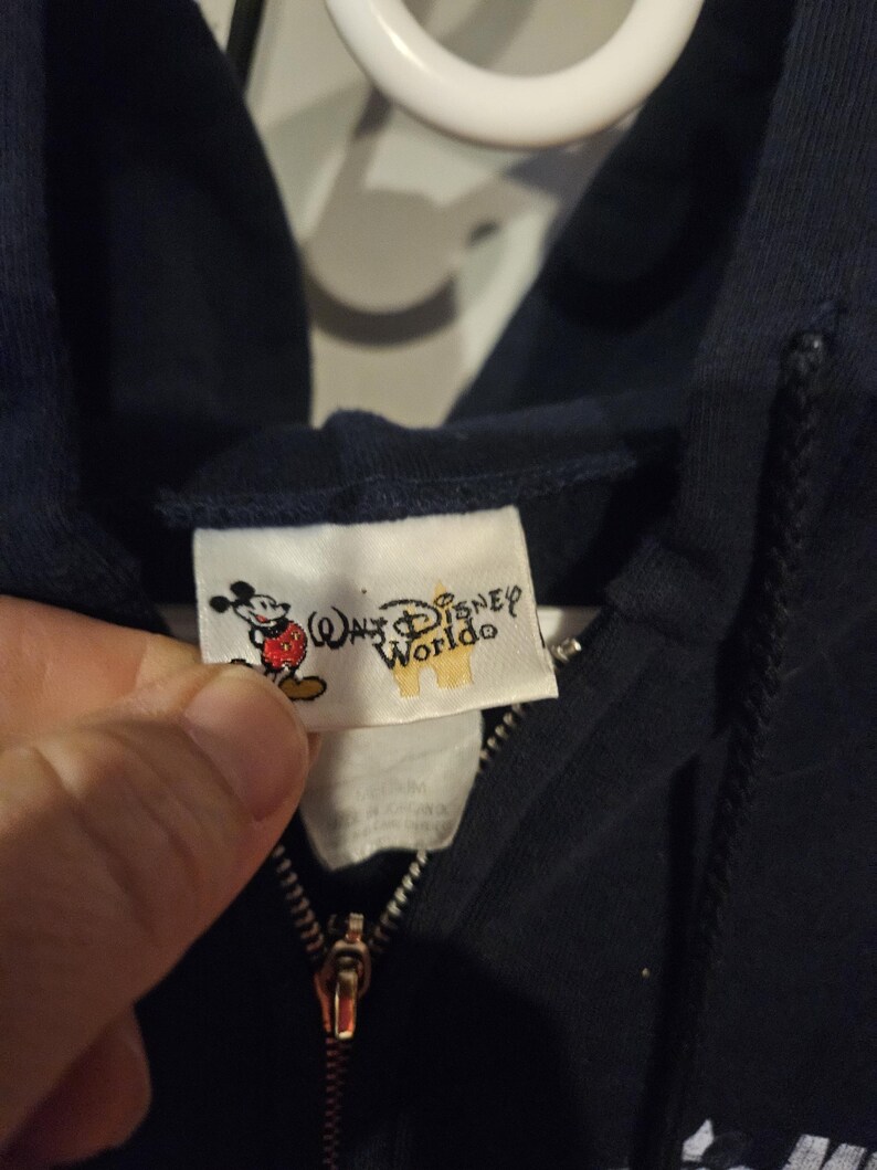 Puede incluir: Sudadera con capucha azul marino con cremallera y una etiqueta con Mickey Mouse y las palabras "Walt Disney World". La cremallera es dorada. La sudadera est&aacute; hecha de un material suave.