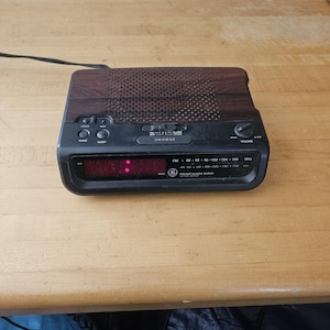 Puede incluir: Un radio reloj GE FM/AM negro y marrón con una pantalla digital. El radio reloj tiene botones para despertar, dormir y posponer. La parte superior de la radio tiene un patrón de vetas de madera.