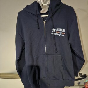Puede incluir: Sudadera con capucha azul marino con cremallera y bolsillo delantero. La sudadera tiene el texto "MICKEY" y "Walt Disney World" en letras blancas y rojas. La cremallera es dorada.