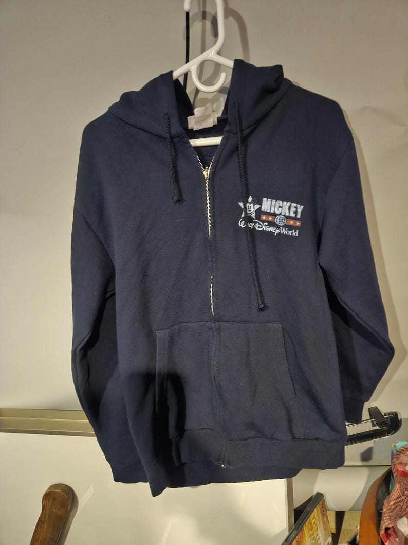 Puede incluir: Sudadera con capucha azul marino con cremallera y bolsillo delantero. La sudadera tiene el texto "MICKEY" y "Walt Disney World" en letras blancas y rojas. La cremallera es dorada.