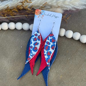 May include: A pair of layered teardrop-shaped earrings featuring a navy blue textured base, a solid red middle layer, and a white top layer with a blue and red leopard print pattern. The earrings are attached to a white display card labeled The Jeweled Hippo.