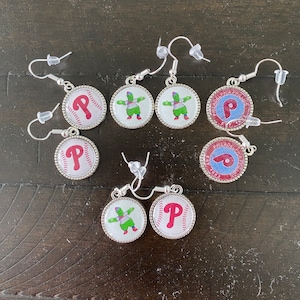 May include: Silver-toned dangle earrings featuring various designs. Some earrings display a red "P" logo on a baseball, while others showcase a green mascot figure. The earrings have a round shape with a decorative border.