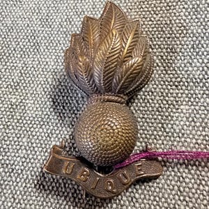 May include: A bronze-coloured metal military badge featuring a flaming grenade design. The spherical base has a textured surface, topped with stylised flames. A curved banner at the bottom displays the embossed text UBIQUE in capital letters.