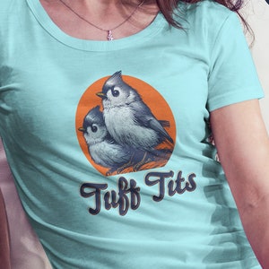 May include: A light blue t-shirt with a graphic of two birds on a branch with a sunset behind them. The text "Tuff Tits" is printed in a retro font below the birds.
