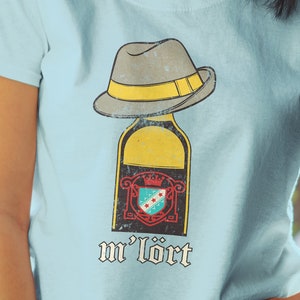 May include: A light blue t-shirt featuring a vintage-style graphic of a bottle wearing a fedora hat. The bottle has a label with a crest and the text "m'lort".