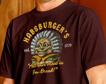 May include: A brown t-shirt with a vintage-style logo for a restaurant called "Habsburger's" established in 1270. The logo features a cartoon burger with a face and a wig. The text below the logo reads "All our meat is In-Bread!"