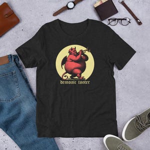 May include: Dark gray t-shirt featuring a red devil playing a trumpet inside a yellow circle. The text "demonic tooter" is below the illustration. The shirt is made of a soft material.