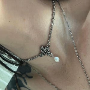 Puede incluir: Collar plateado con diseño floral y perla colgante. El collar tiene una cadena delicada y un pendiente a juego con una perla. La joya está diseñada para llevarse alrededor del cuello y la oreja.
