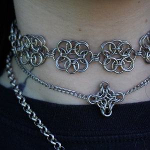 Puede incluir: Un collar choker de malla de plata con un colgante de malla de plata que cuelga debajo. El collar está superpuesto a otras dos cadenas de plata.