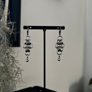 May include: A pair of silver chain earrings with a small ball at the end of each earring. The earrings are hanging from a black stand.
