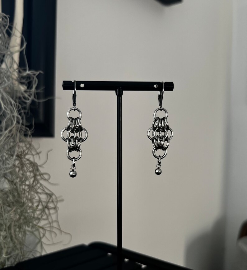 May include: A pair of silver chain earrings with a small ball at the end of each earring. The earrings are hanging from a black stand.