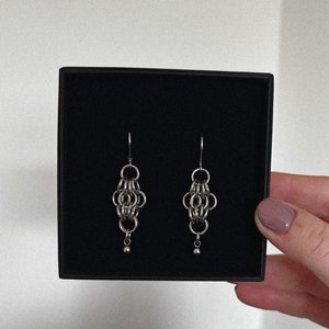 May include: A pair of silver chain earrings with a small ball at the end of each earring. The earrings are displayed in a black box.