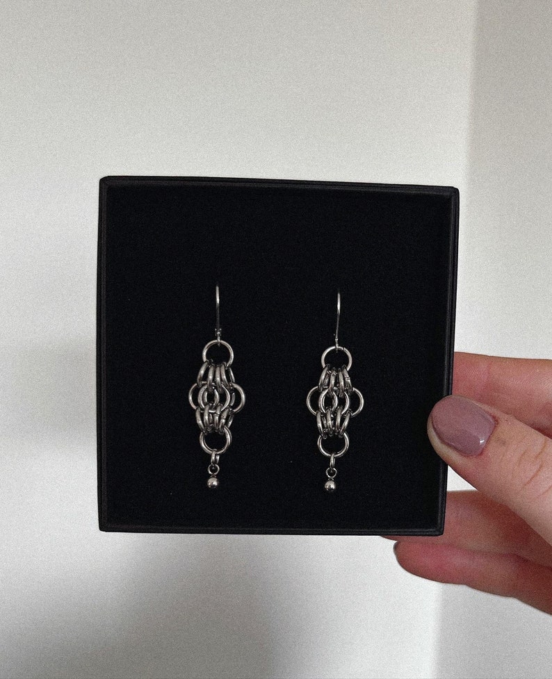May include: A pair of silver chain earrings with a small ball at the end of each earring. The earrings are displayed in a black box.