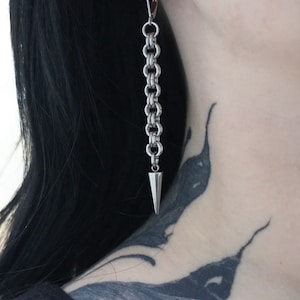 May include: A pair of silver earrings with a long chain and a pointed tip. The earrings are hanging from the earlobes of a person with black hair.